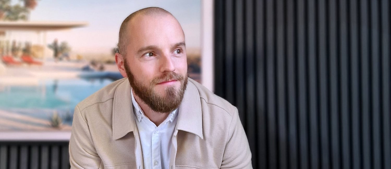 Adam Van den Houten, architect and co-founder of Enza Architects, seated in front of framed artwork in a modern office setting